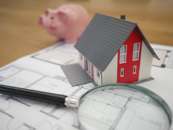 A small plastic model of a house, a magnifying glass and a piggy bank lay upon a floor plan document.