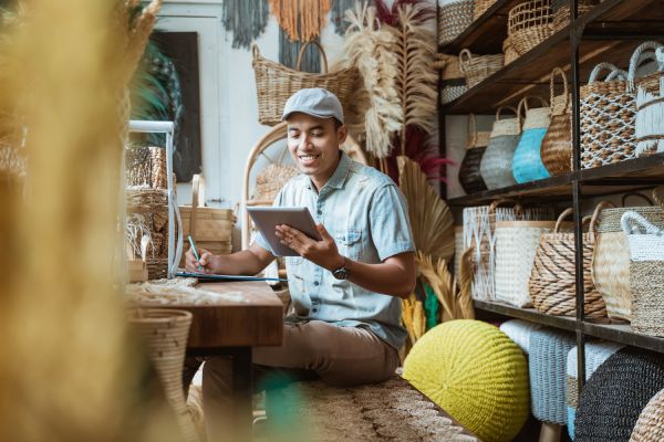 A smiling young business owner sits in his new shop and makes notes whilst researching on his tablet.